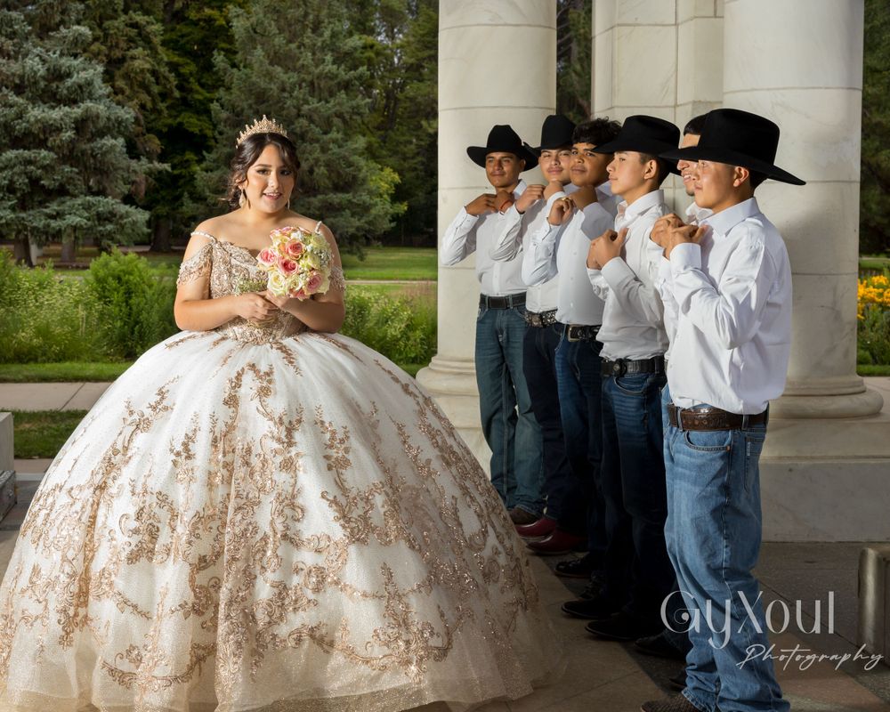 A quinceañera in a gold-embellished gown poses with attendants in matching western wear by white columns.