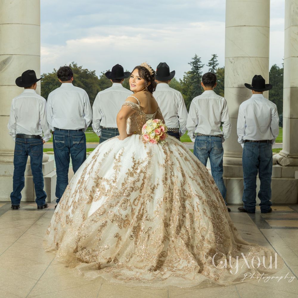 A quinceañera in an elaborately beaded gown poses while attendants face away at a columned venue.