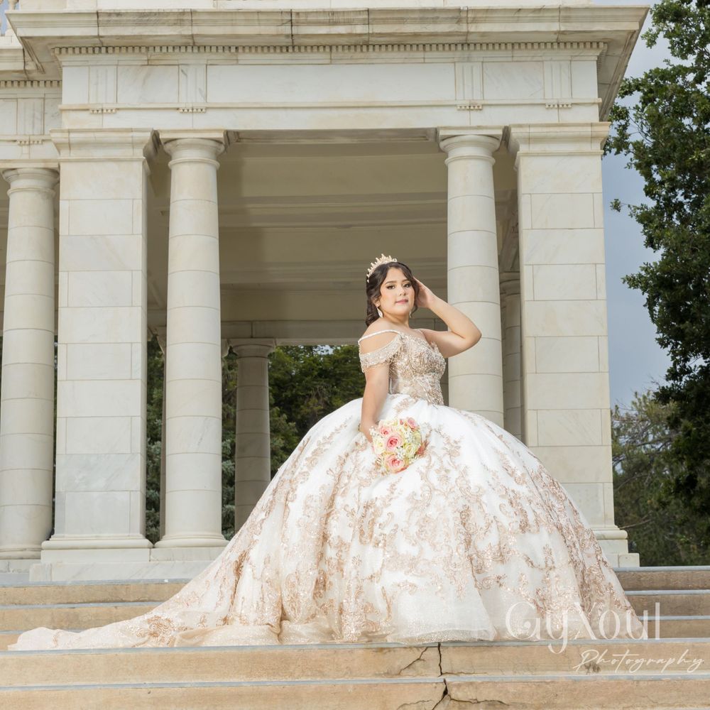 A quinceañera in an ornate champagne-colored ball gown poses elegantly between classical white columns.