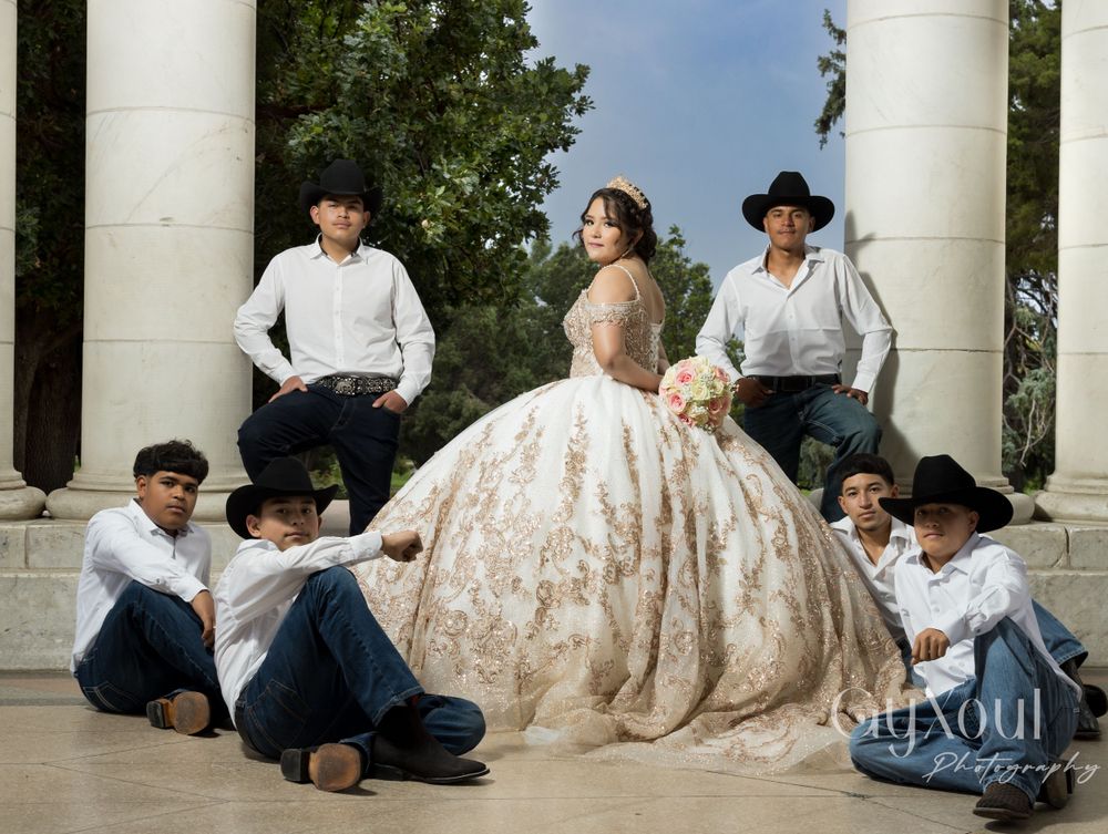 A quinceañera wearing an ornate champagne ball gown poses with attendants in cowboy attire at a columned venue.