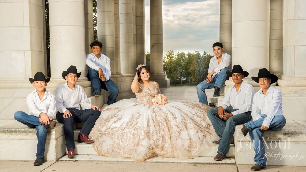 A formal portrait of a quinceañera in a beige ball gown surrounded by attendants at a classical outdoor setting.