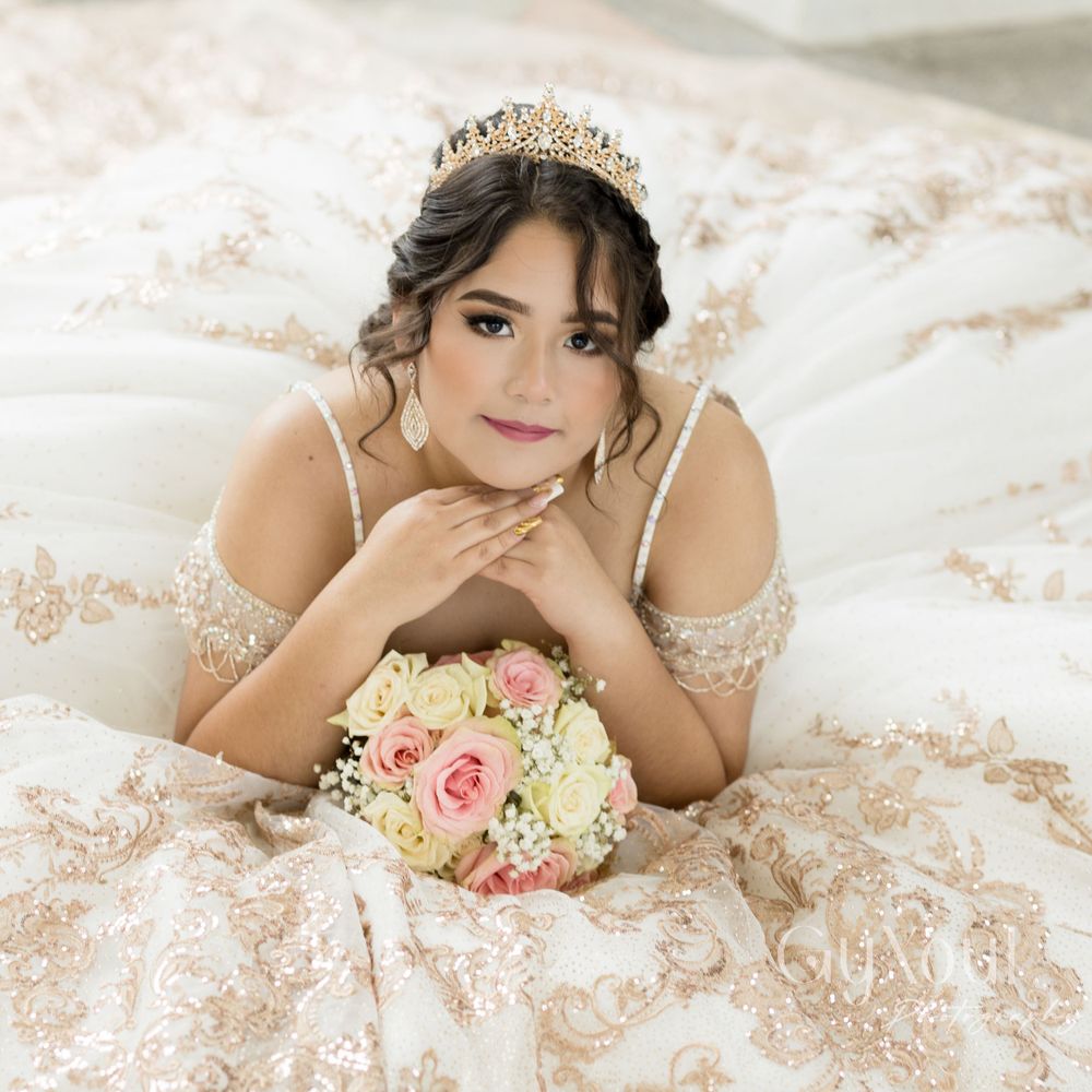 A person in an ornate champagne-colored quinceañera dress with a tiara poses with a pink and white bouquet.