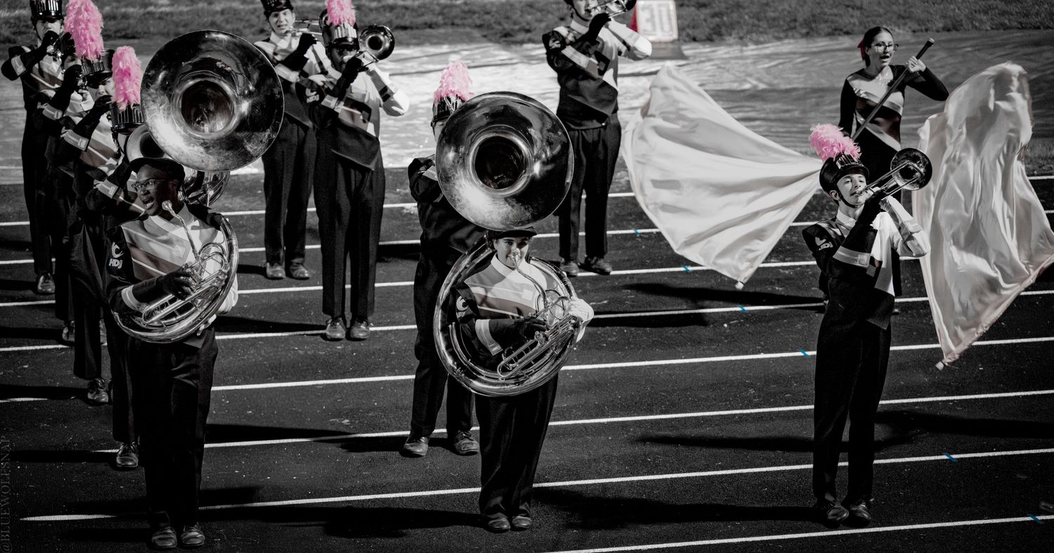 Marching band performers in black uniforms playing tubas and brass instruments on a track field during a night performance.
