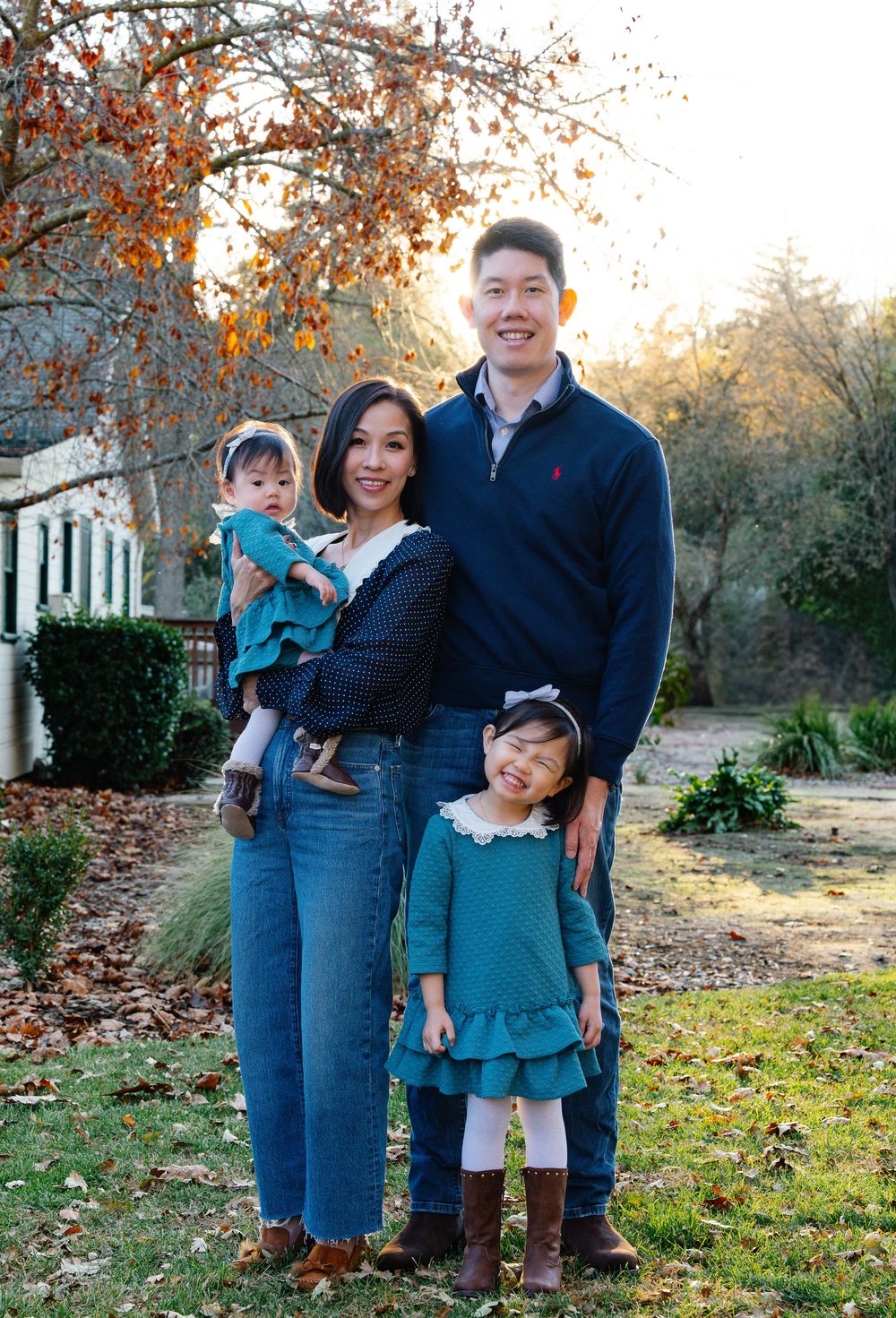 A family stands together outdoors on an autumn day with fallen leaves and trees in the background.
