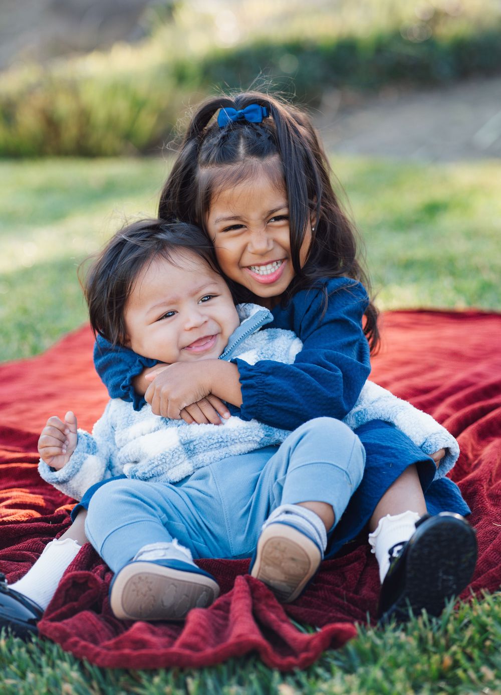 Two children sitting on a red blanket outdoors share a sweet moment of laughter and hugs on a sunny day.