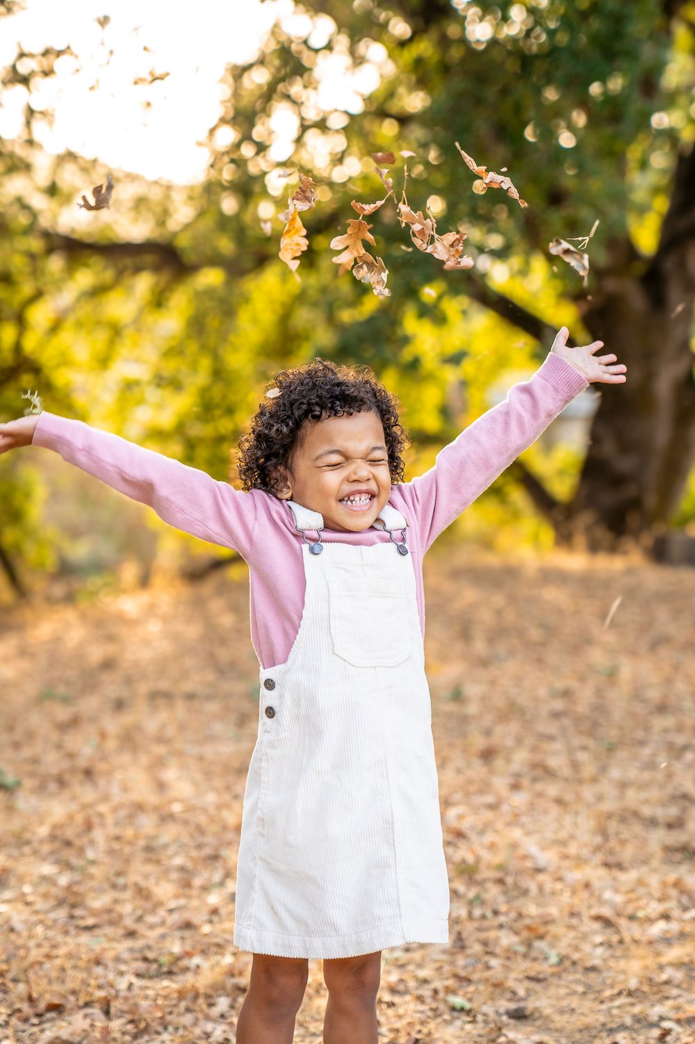 A child in white overalls and pink sweater joyfully throws leaves in golden autumn light.