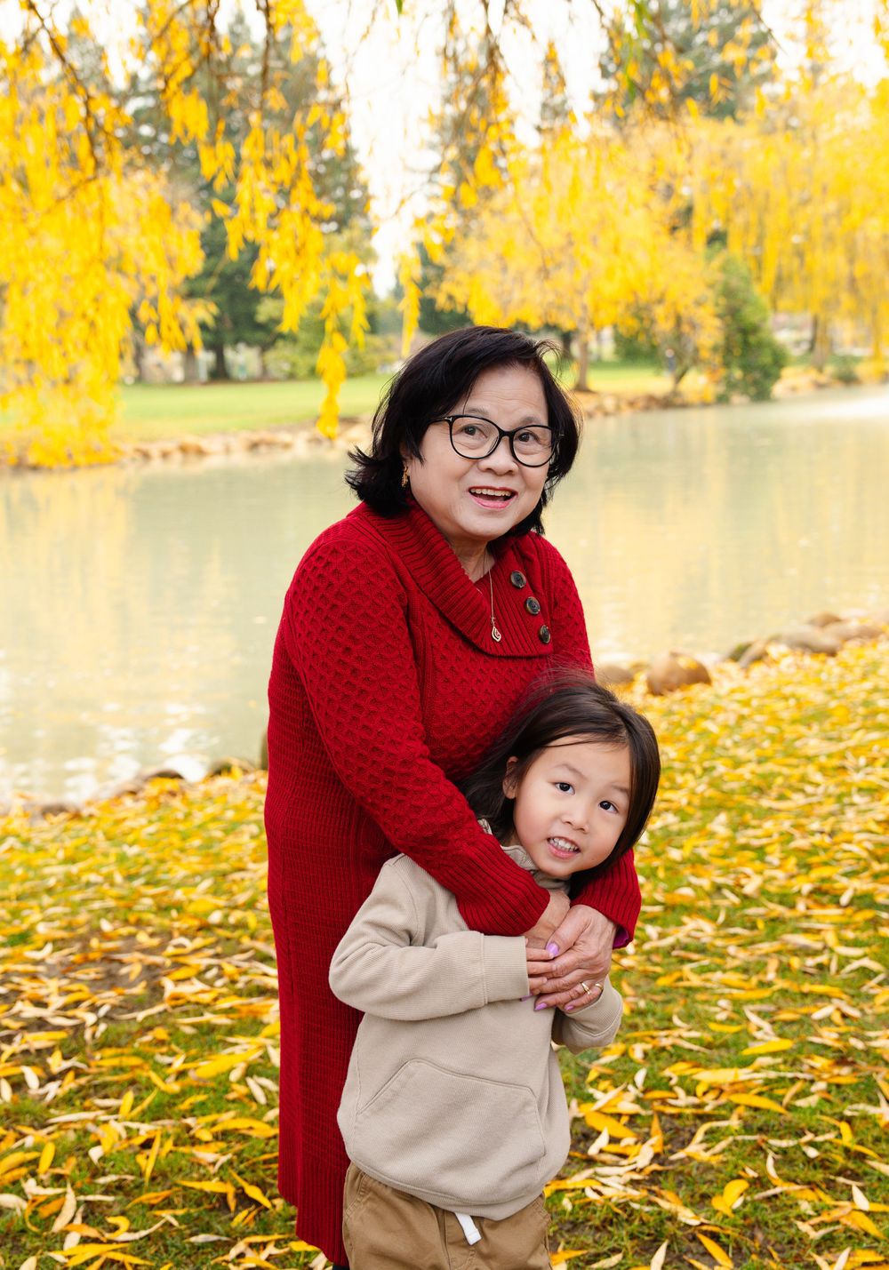 Two people in warm clothing embrace while standing on yellow fallen leaves near a pond on an autumn day.