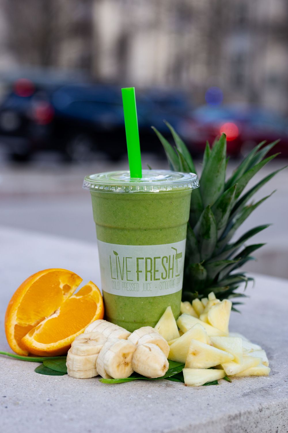 A green smoothie in a clear plastic cup with fresh pineapple, orange slices and banana arranged beside it.