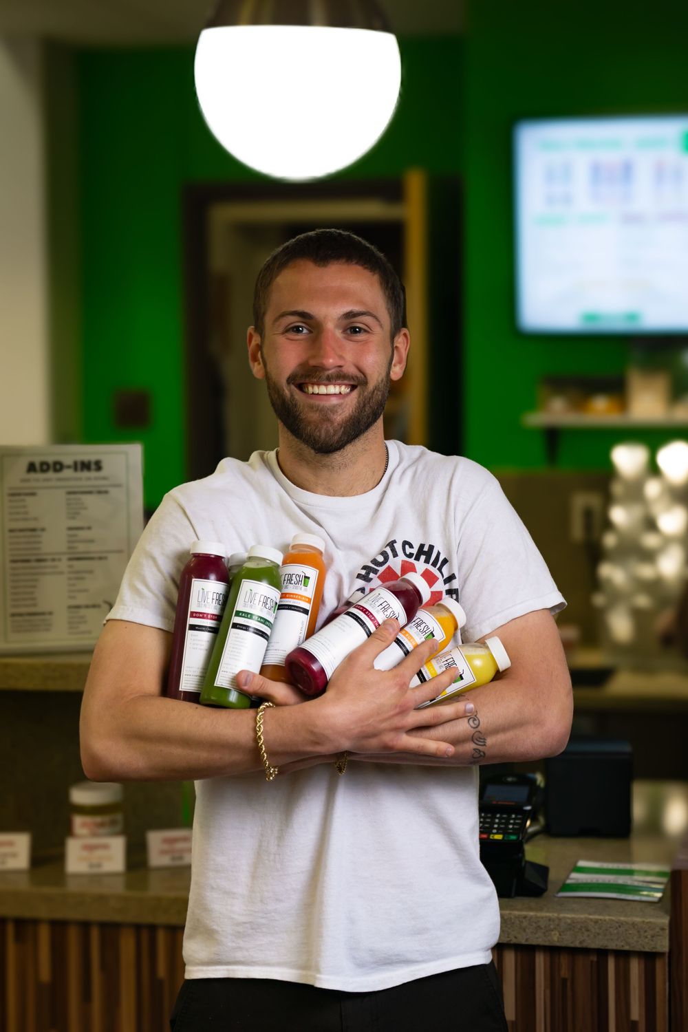 Person in white t-shirt displays colorful juice bottles at a bar counter in green-lit environment.