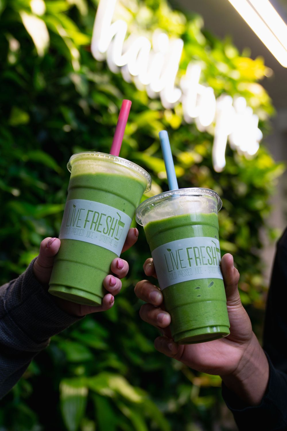 Two green smoothie drinks in clear plastic cups with straws are held up against a leafy green background.