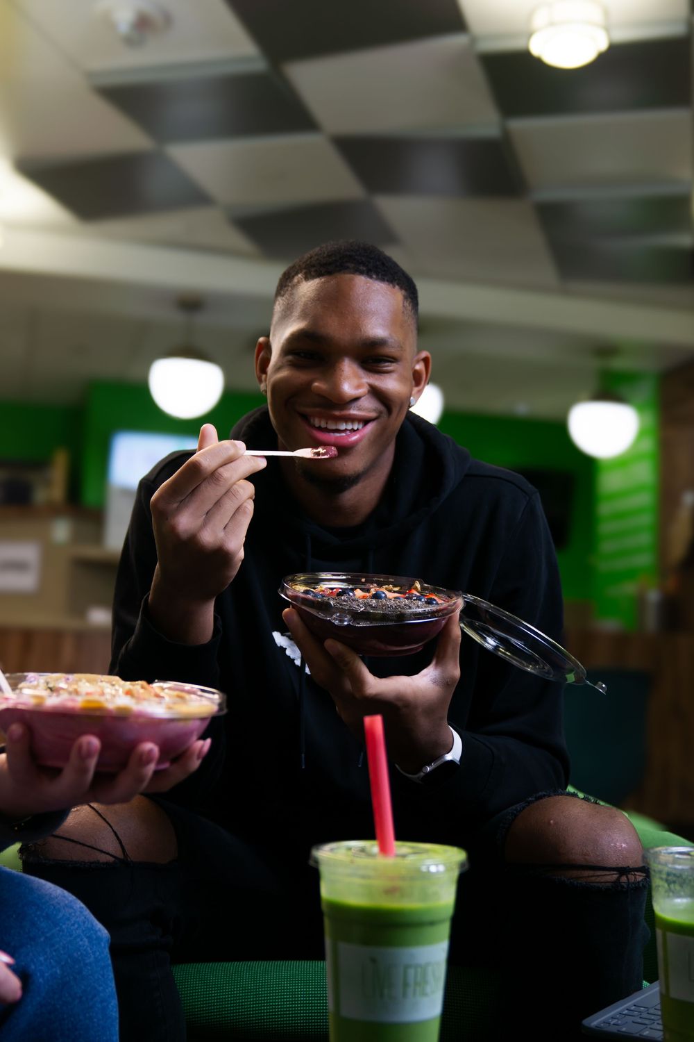 A person enjoys a series of colorful drinks and desserts at a cafe with green lighting in the background.