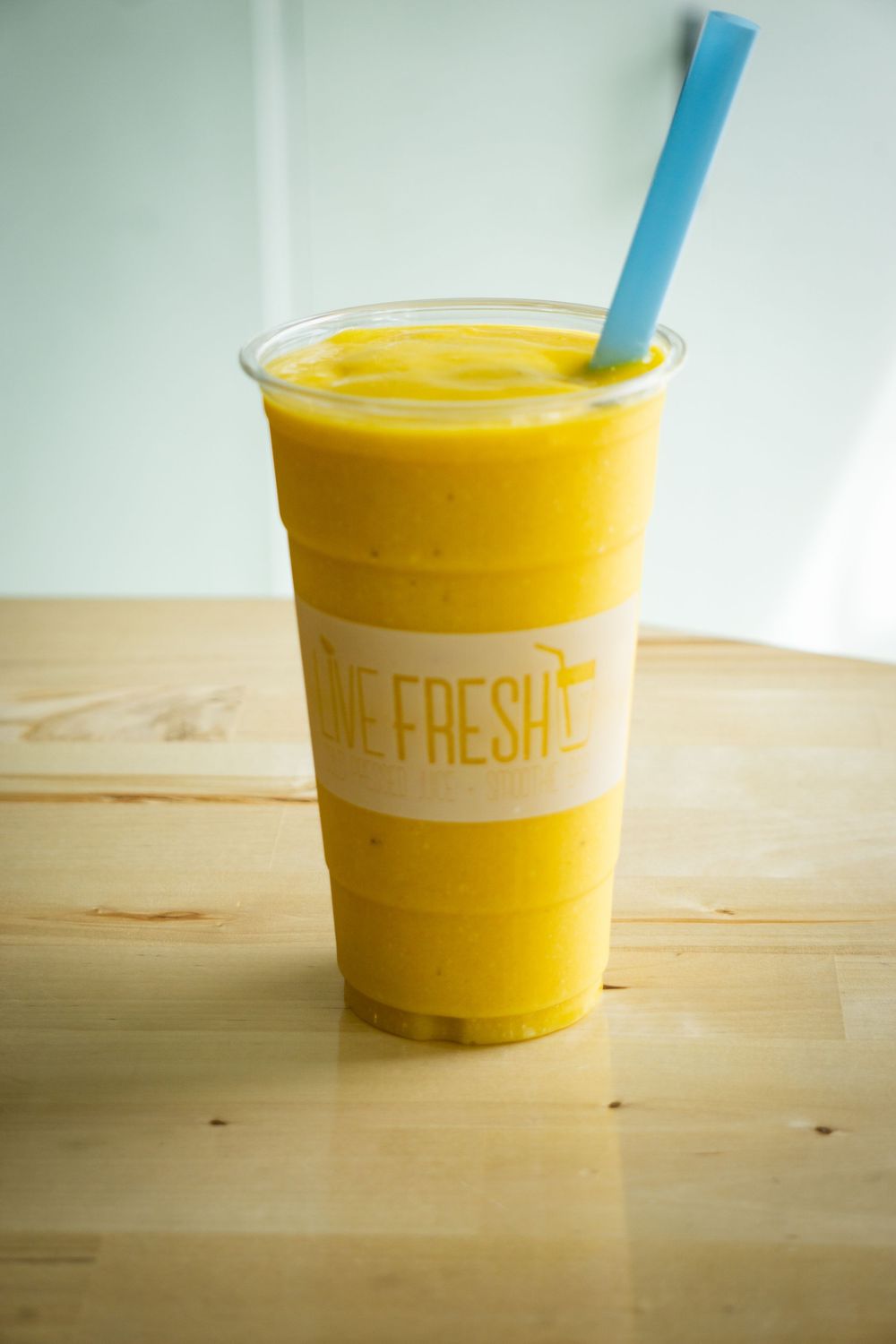 A vibrant yellow smoothie in a clear plastic cup with a blue straw sits on a wooden surface.