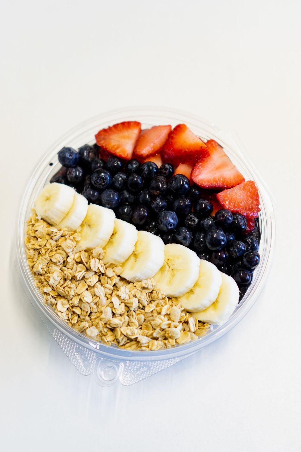 Fresh fruit bowl with sliced bananas, strawberries, blueberries and granola arranged in neat rows on a white plate.