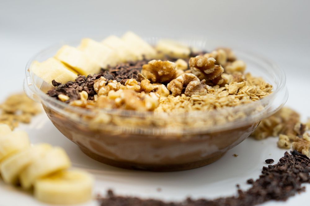 A chocolate smoothie bowl topped with sliced bananas, granola, nuts and chocolate shavings on a light background.
