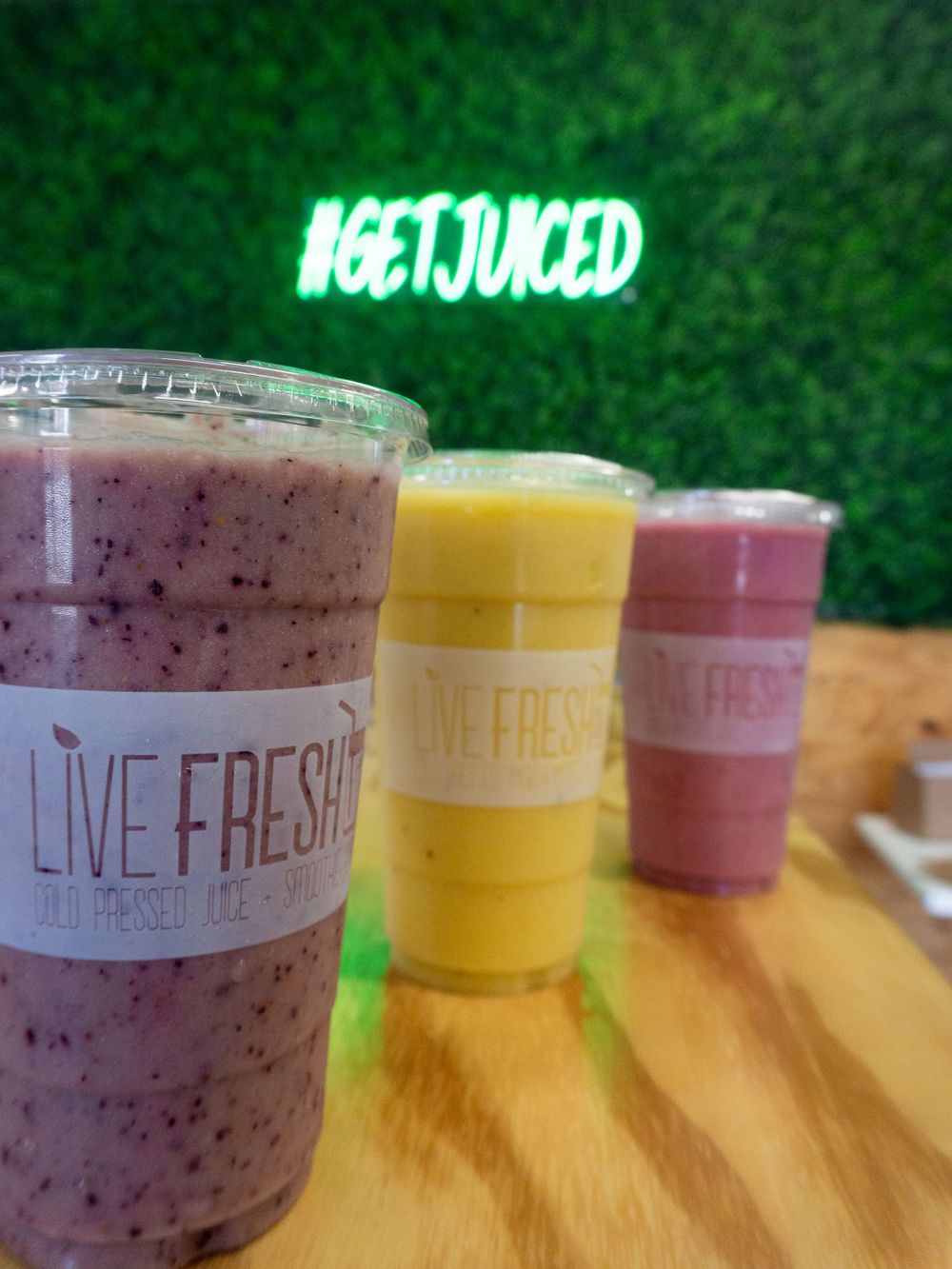 Colorful smoothie drinks in clear plastic cups arranged in a row on a wooden surface with green foliage background.