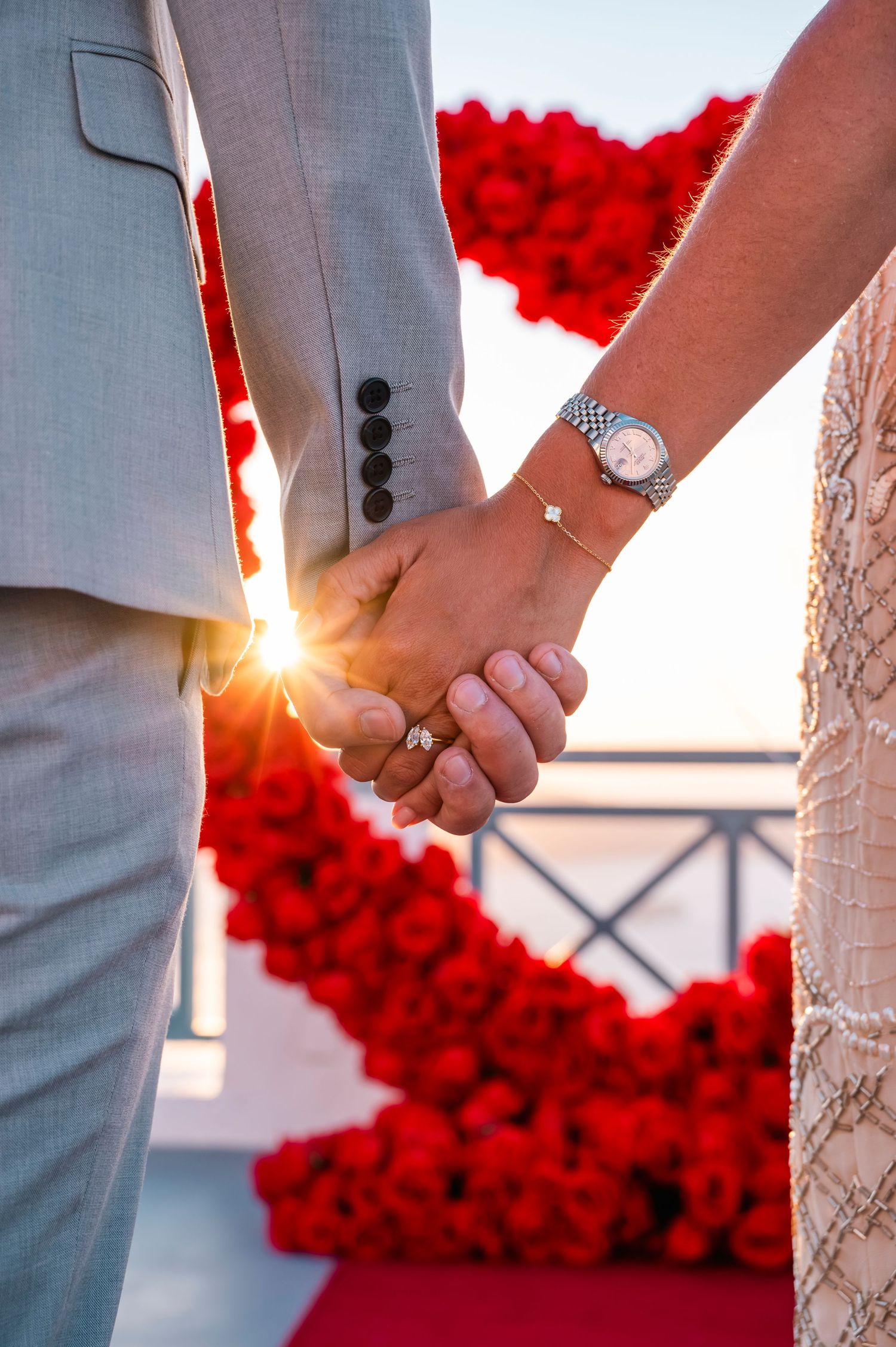 Close-up of engaged couple holding hands with red rose backdrop and sunlight filtering through.