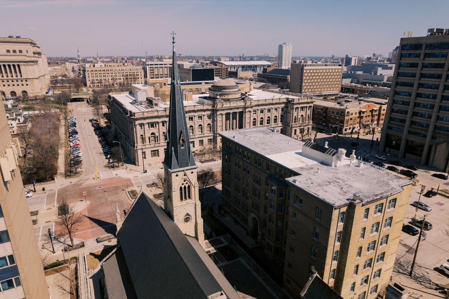 Aerial view of a historic church spire and urban buildings in downtown Milwaukee with the city skyline in the distance.