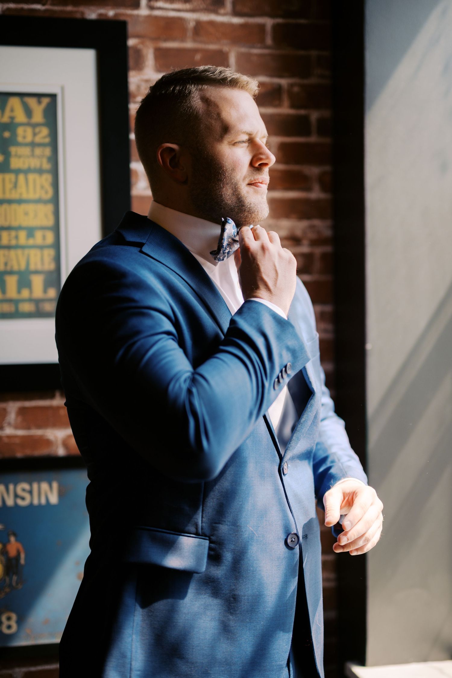 A person in a navy blue suit adjusts their bow tie while standing near a brick wall with vintage posters.