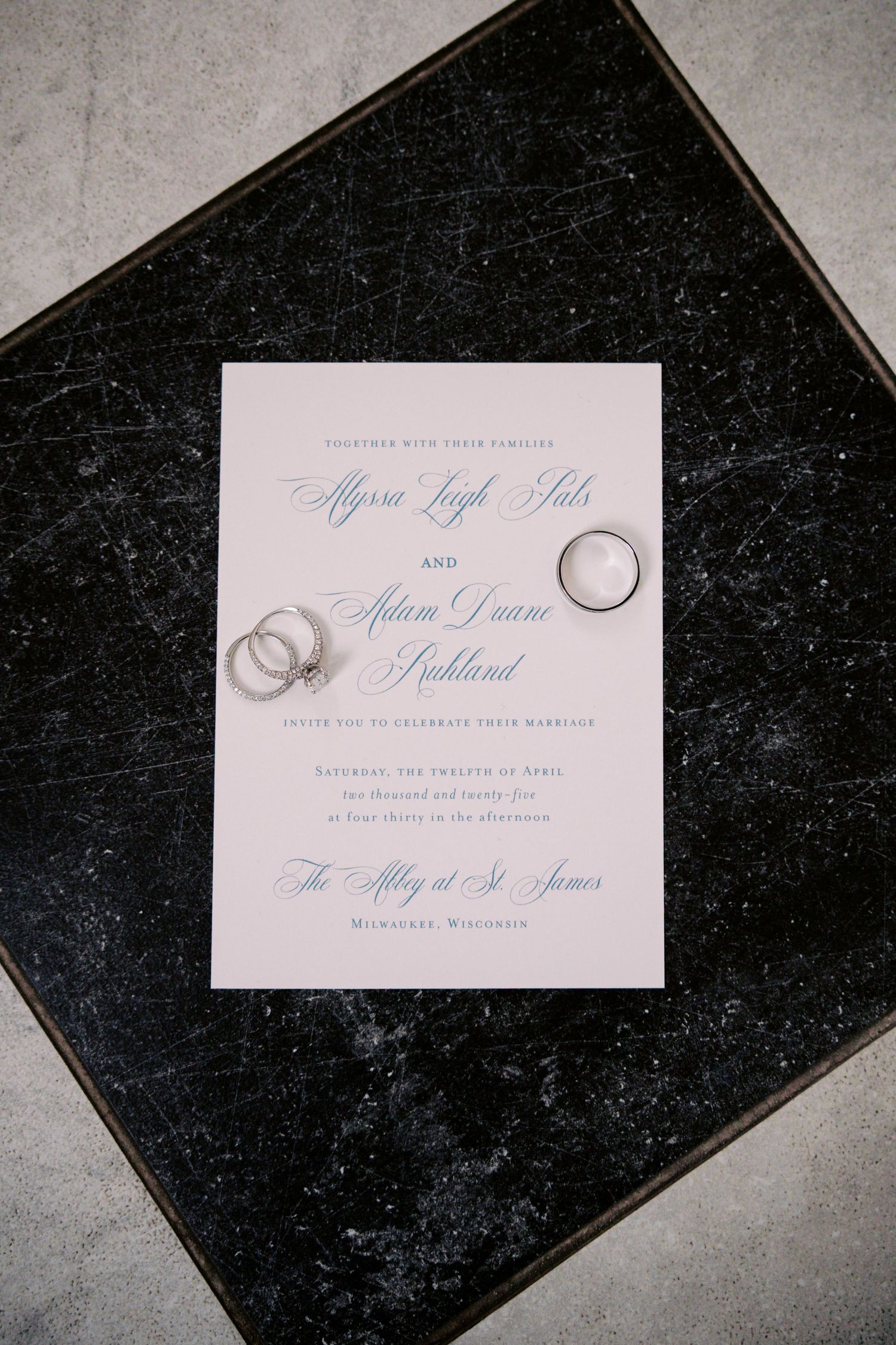 Wedding invitation with elegant blue calligraphy text and silver wedding bands on a black background surface.