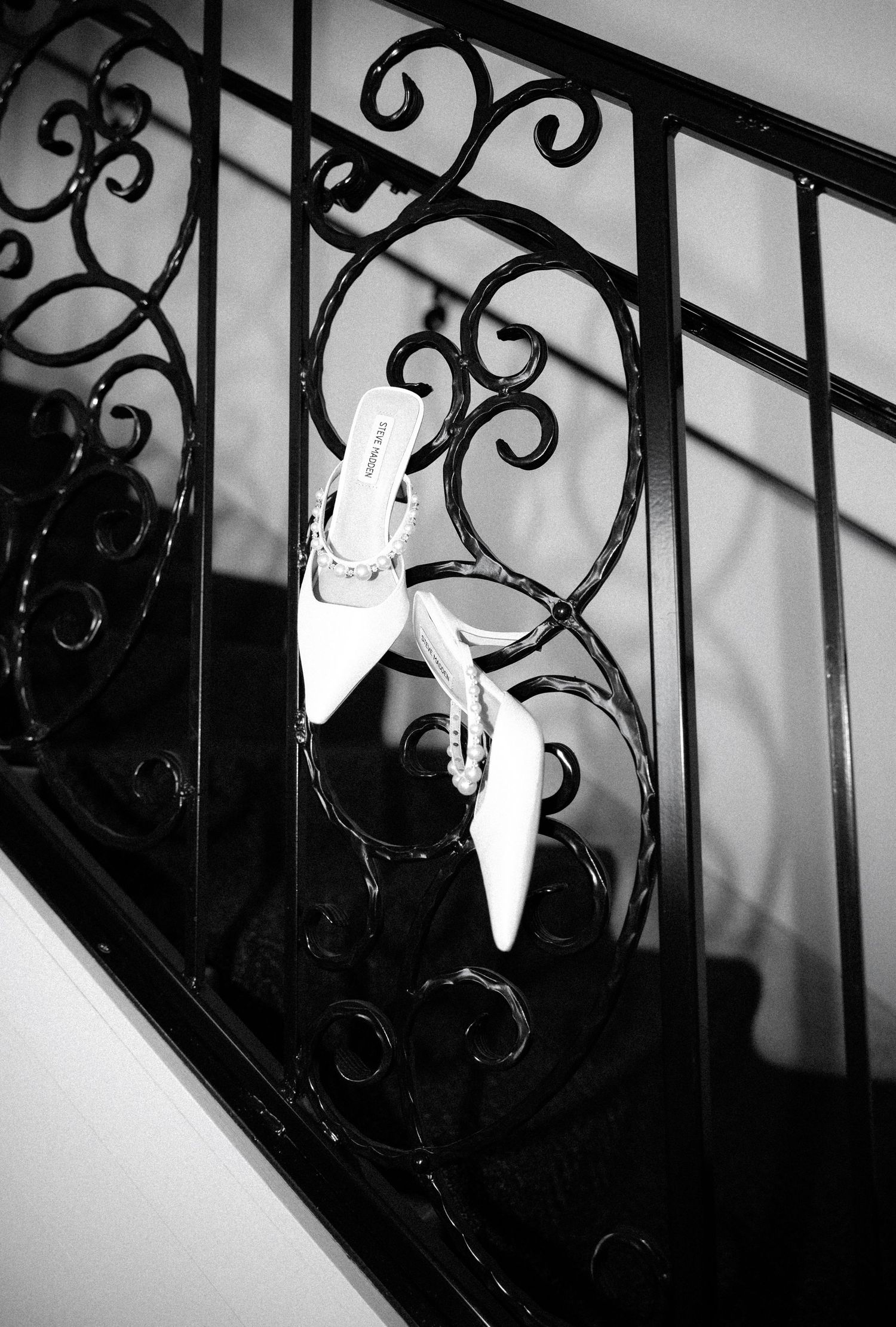 Black and white photo of white high heel shoes perched on an ornate wrought iron staircase railing with decorative scrolls.