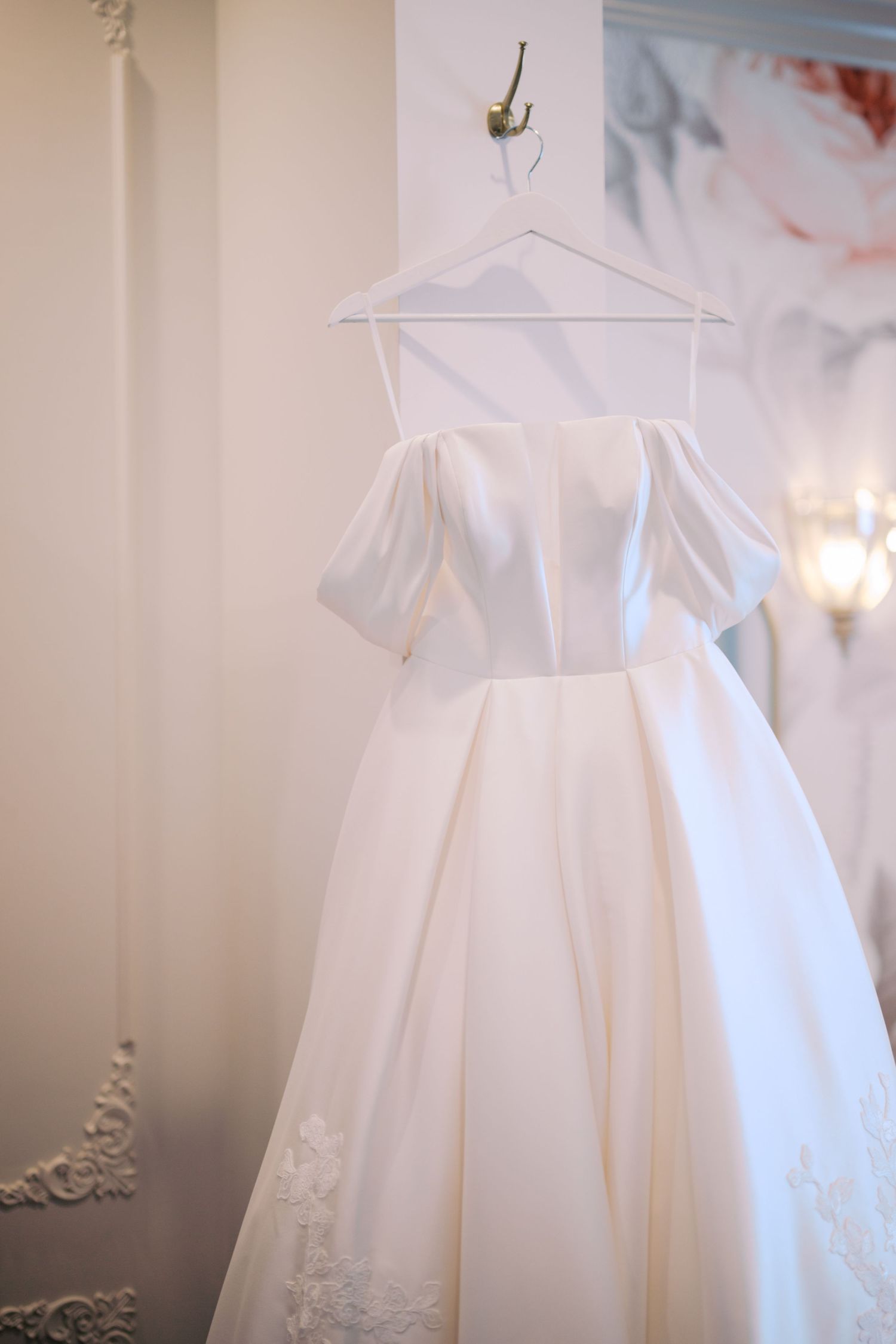 Elegant white wedding dress with off-shoulder details and full pleated skirt hanging on display against light wall.