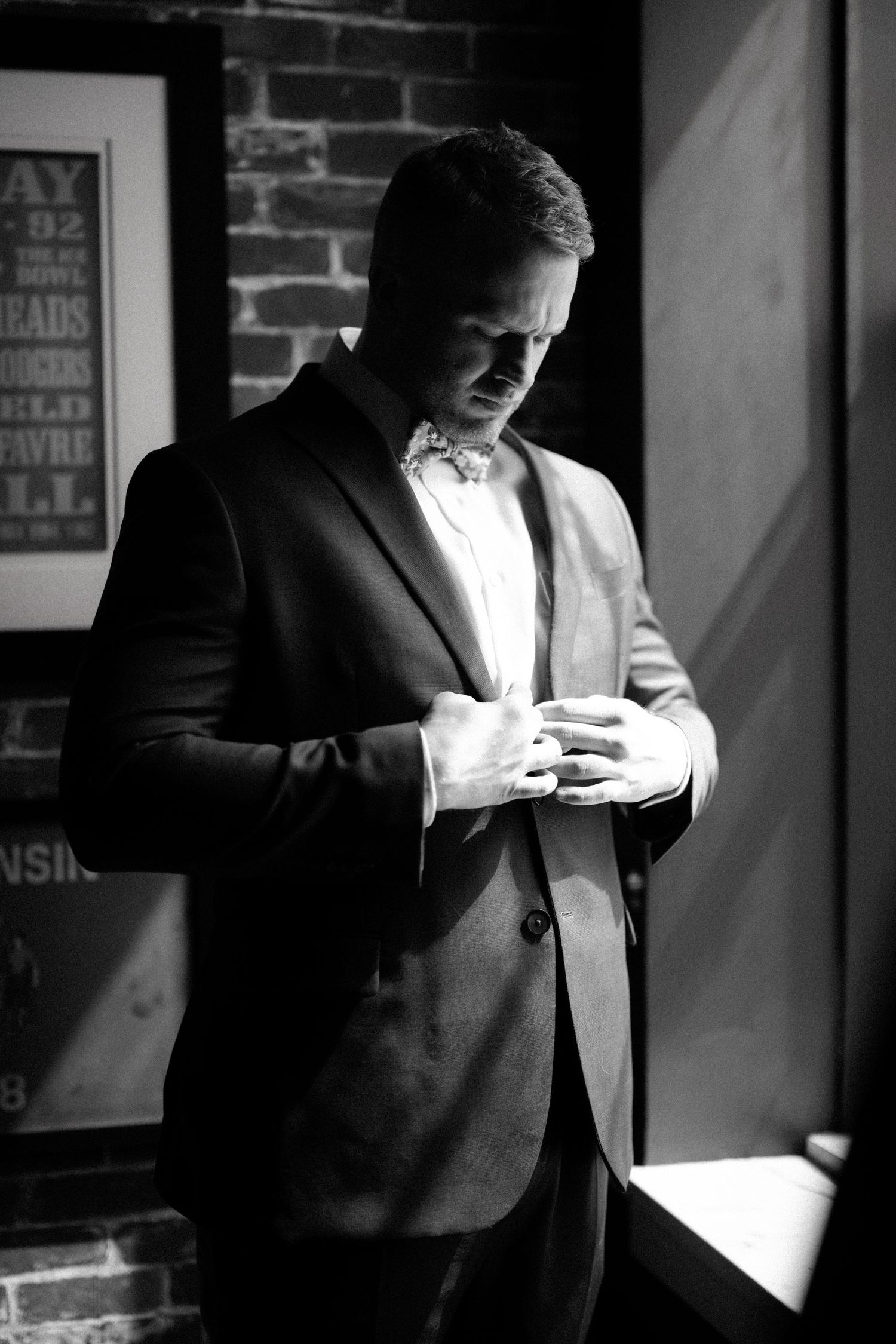 A person in a suit and bowtie stands by a brick wall in dramatic black and white lighting while checking their phone.