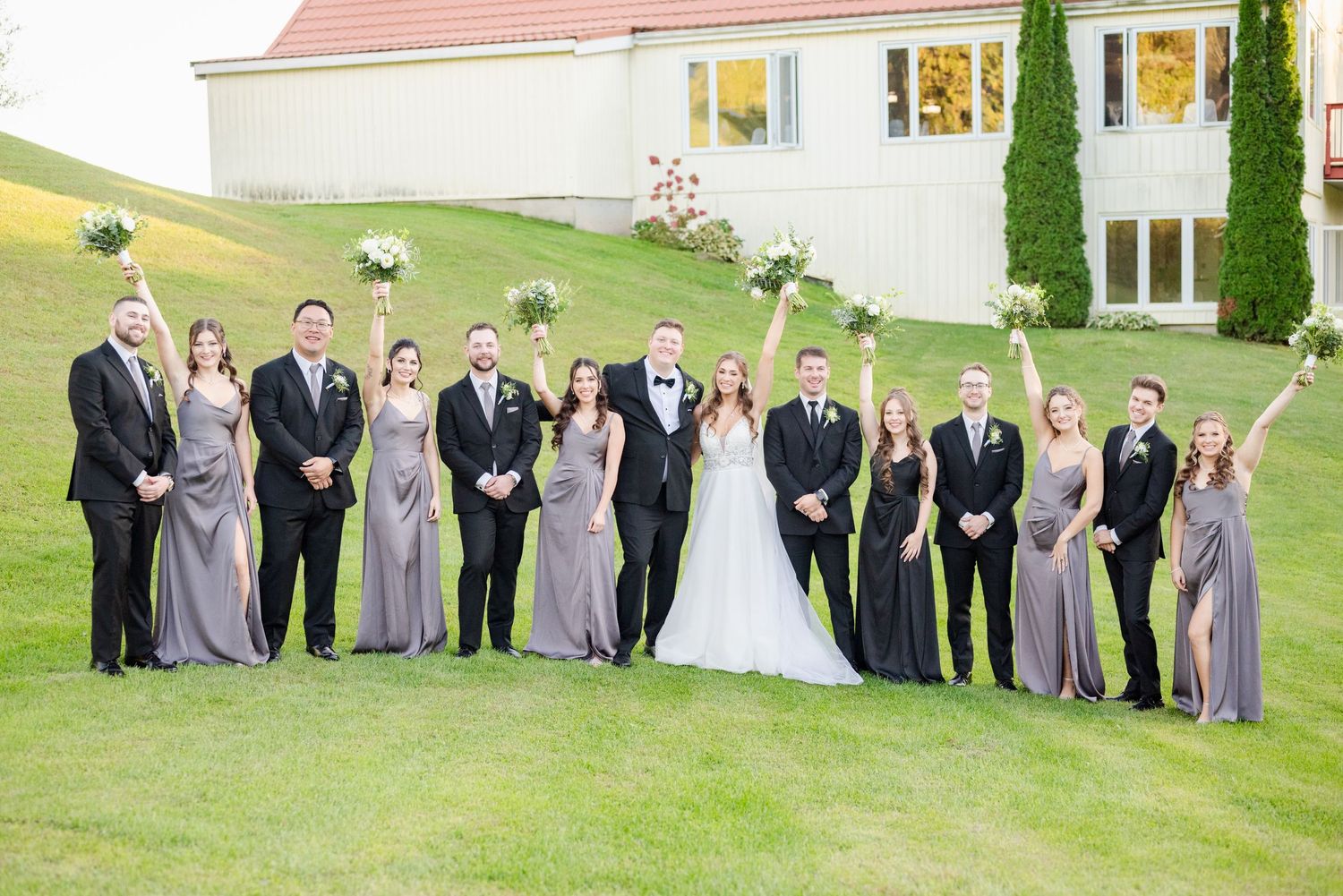 Wedding party in formal attire poses on green lawn with white building and cypress trees in background.