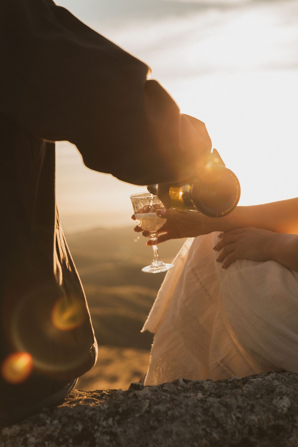 Two people holding wine glasses toast at sunset with golden light filtering through creating a romantic silhouette.