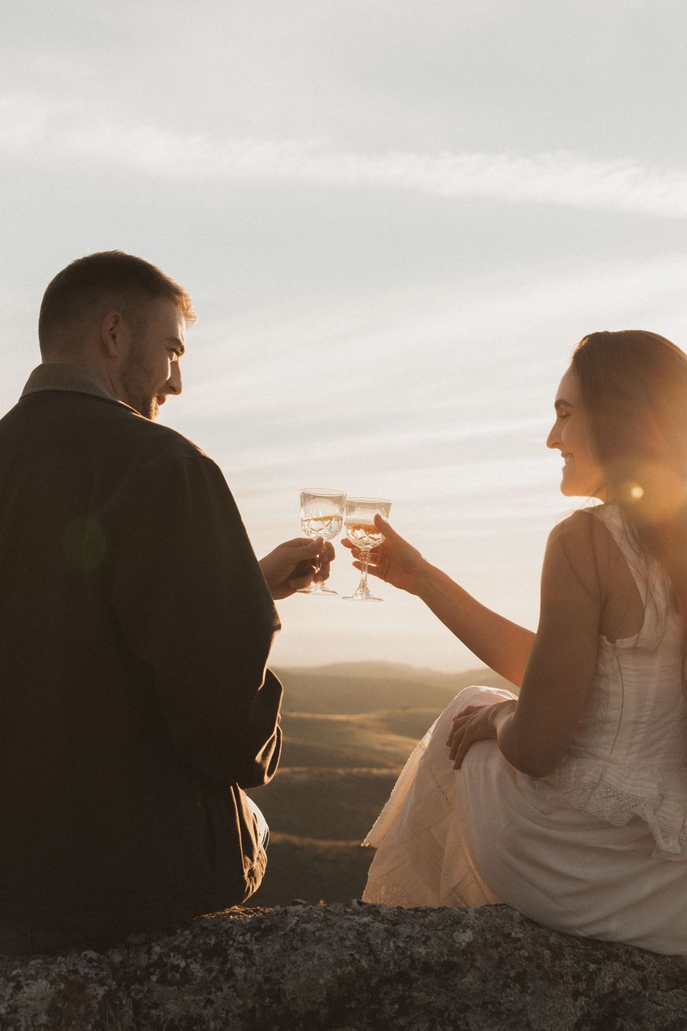 Silhouetted couple toasts wine glasses against a golden sunset on a hilltop during a romantic evening.
