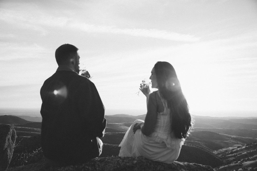 Two people sit together silently watching a scenic mountain vista at sunset from a high viewpoint.