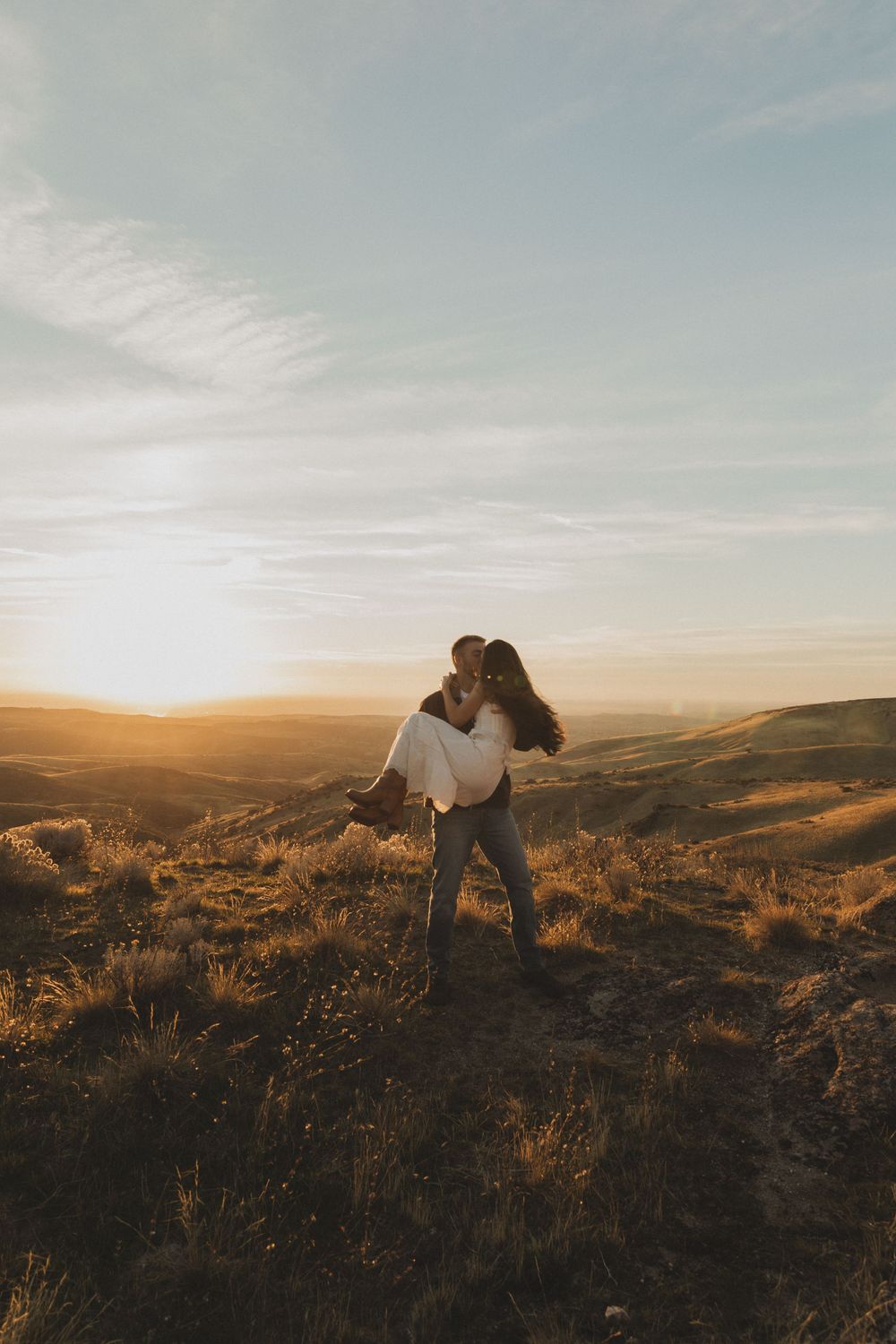 A couple embraces on a hilltop during golden hour with a sweeping desert landscape and mountains in the background.