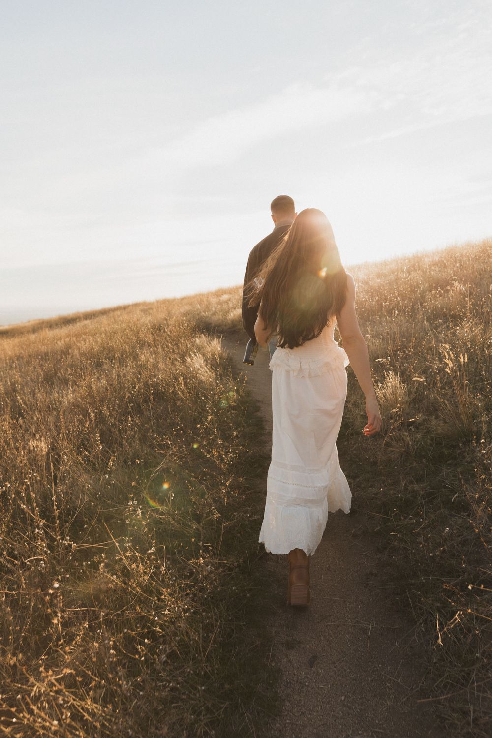 A couple walks hand in hand along a dirt path through golden grass at sunset.