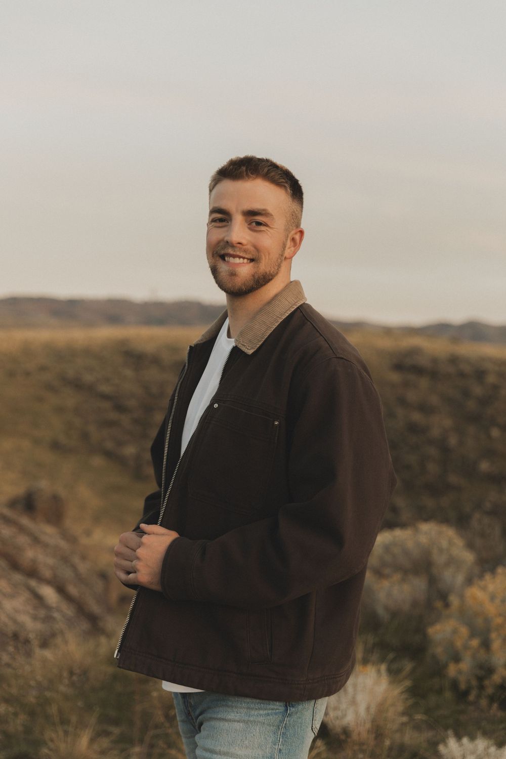 Person in a brown jacket and jeans stands against a desert landscape at golden hour.