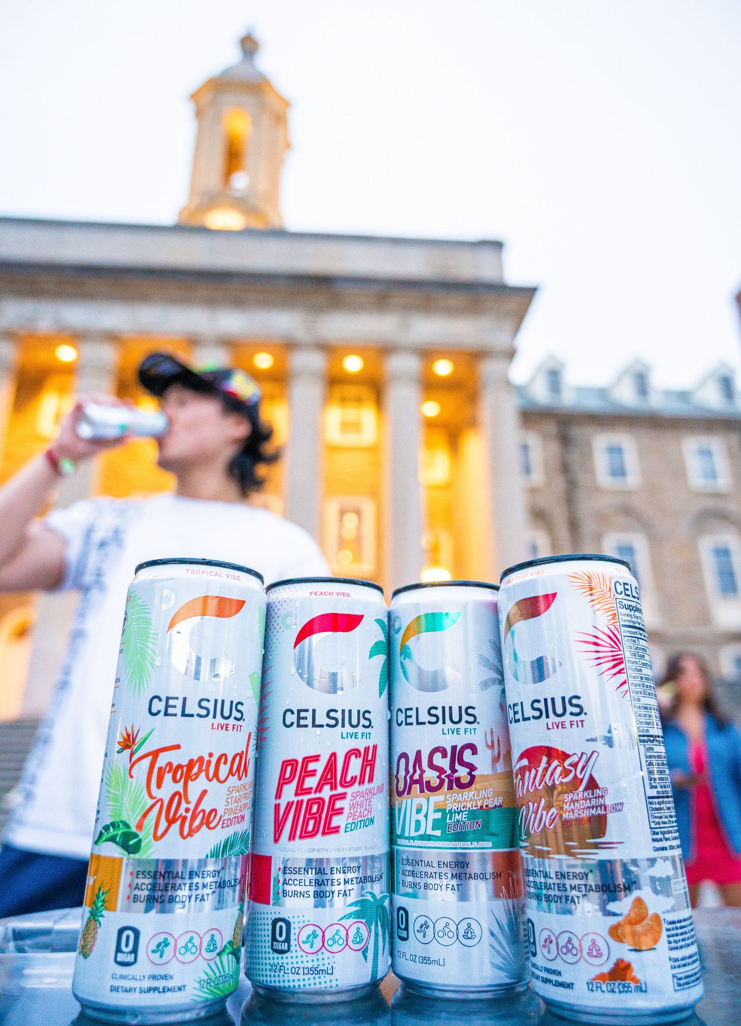 Four cans of Celsius energy drink flavors displayed in front of an illuminated classical building at dusk.