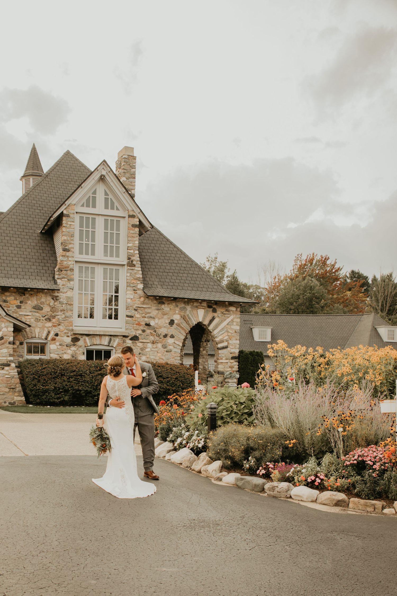 A couple stands together outside a stone cottage with tall windows and colorful garden beds along the driveway.