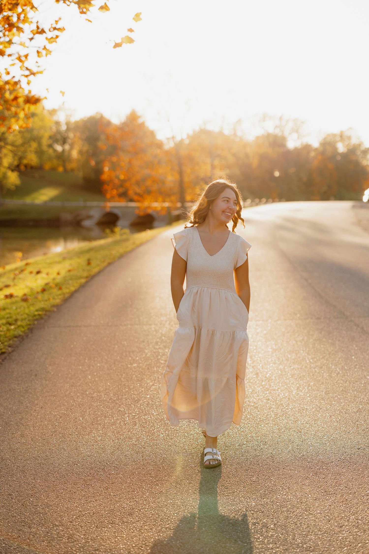 Someone in a flowing white dress walks down a sunlit road during golden hour with autumn trees in the background.