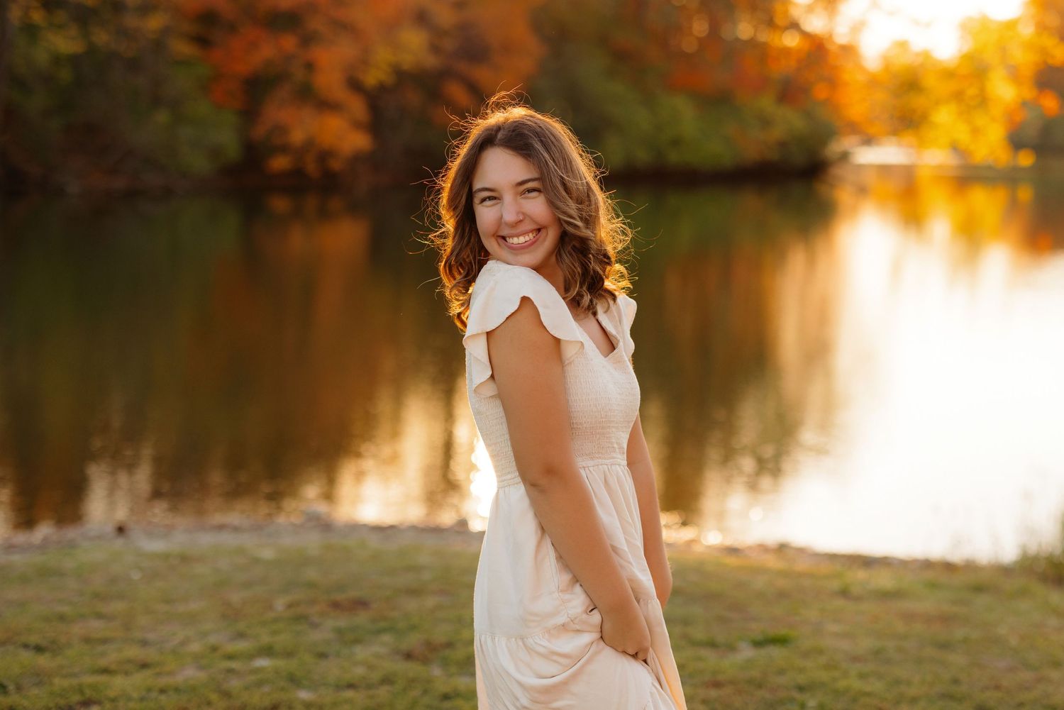 Person in white dress poses by a lake at sunset with warm autumn colors reflecting on the water.