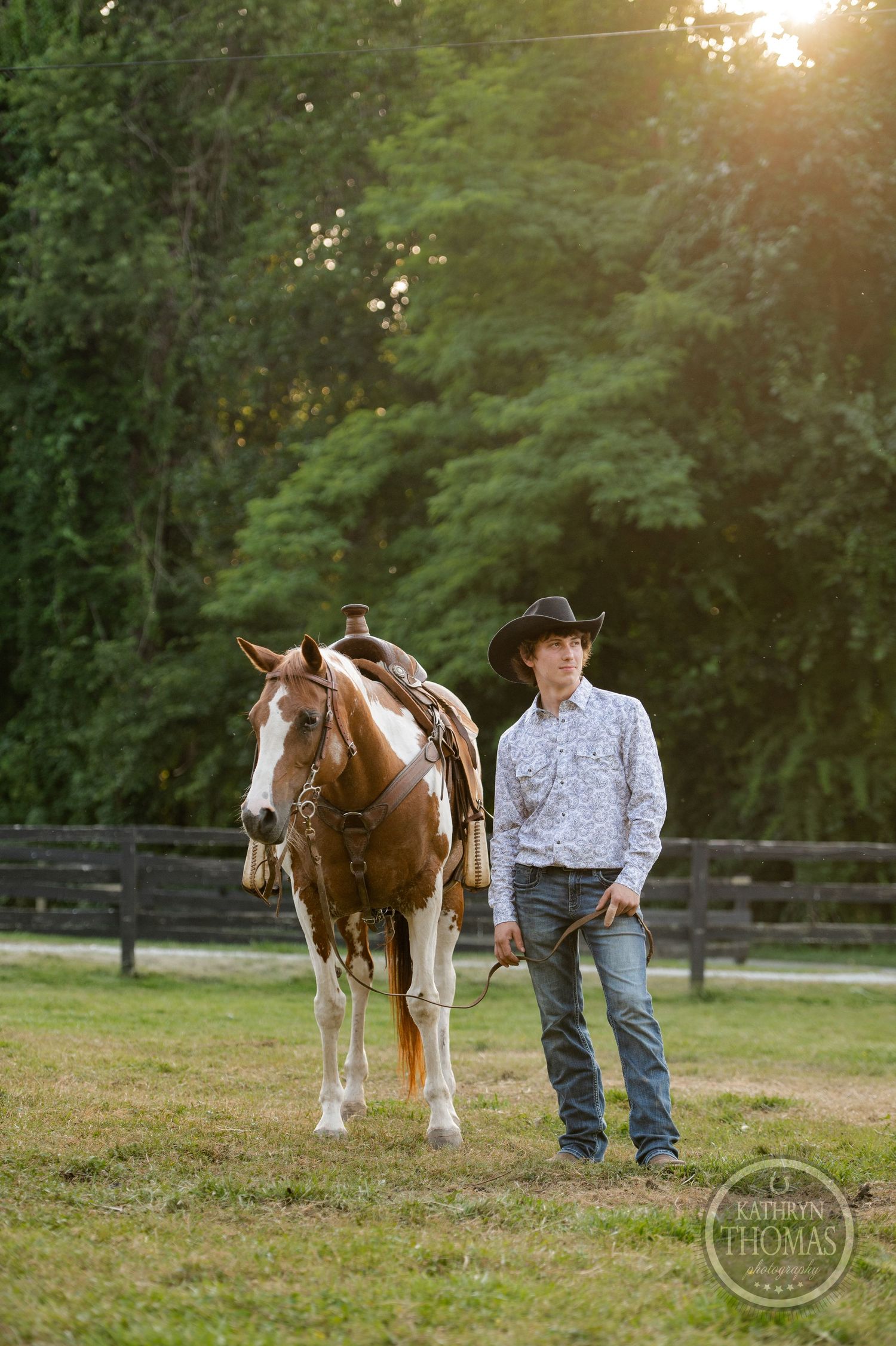 A painted horse with western tack stands beside someone in a fenced field at dusk.