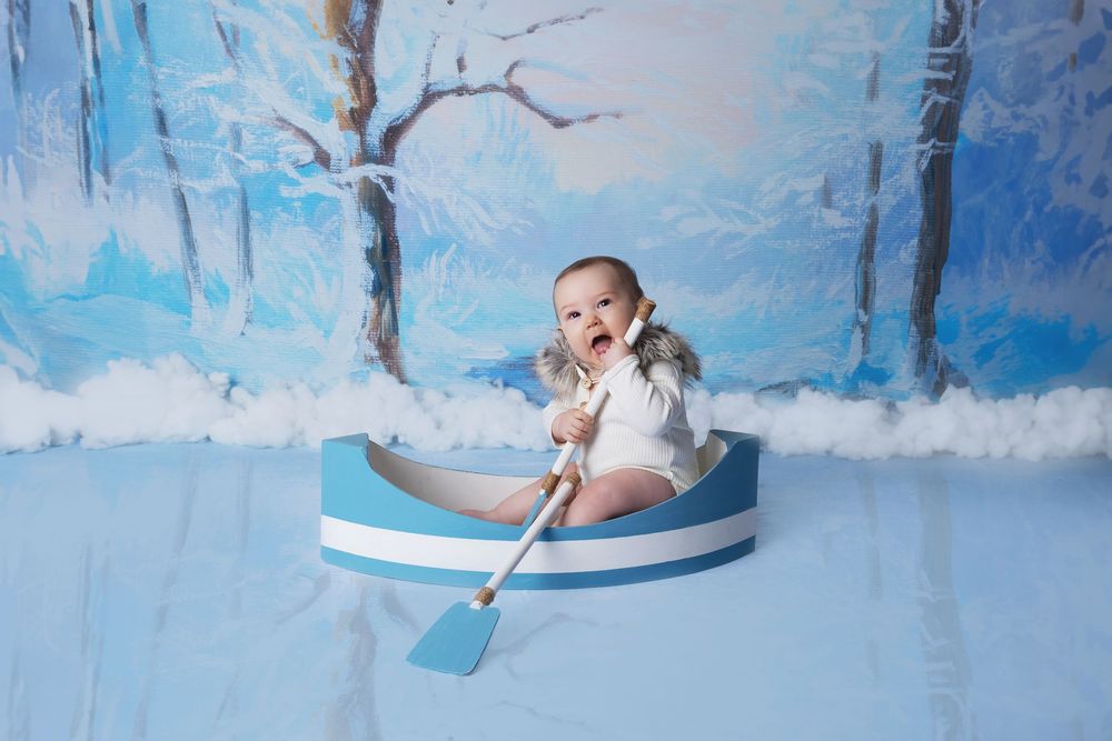 Small child in winter outfit sits in blue boat prop against snowy winter backdrop for studio photography.