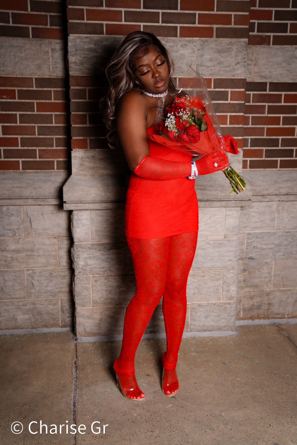 Someone in a striking red dress and matching tights poses with red roses against a brick and stone wall.