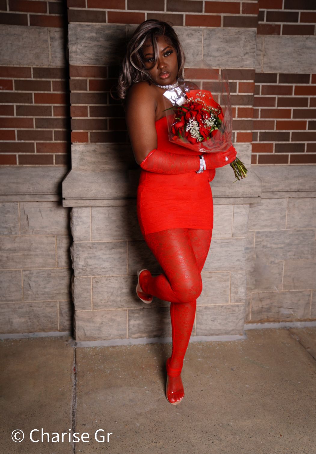A figure in a striking red dress and matching tights poses against a brick wall while holding a vibrant floral bouquet.