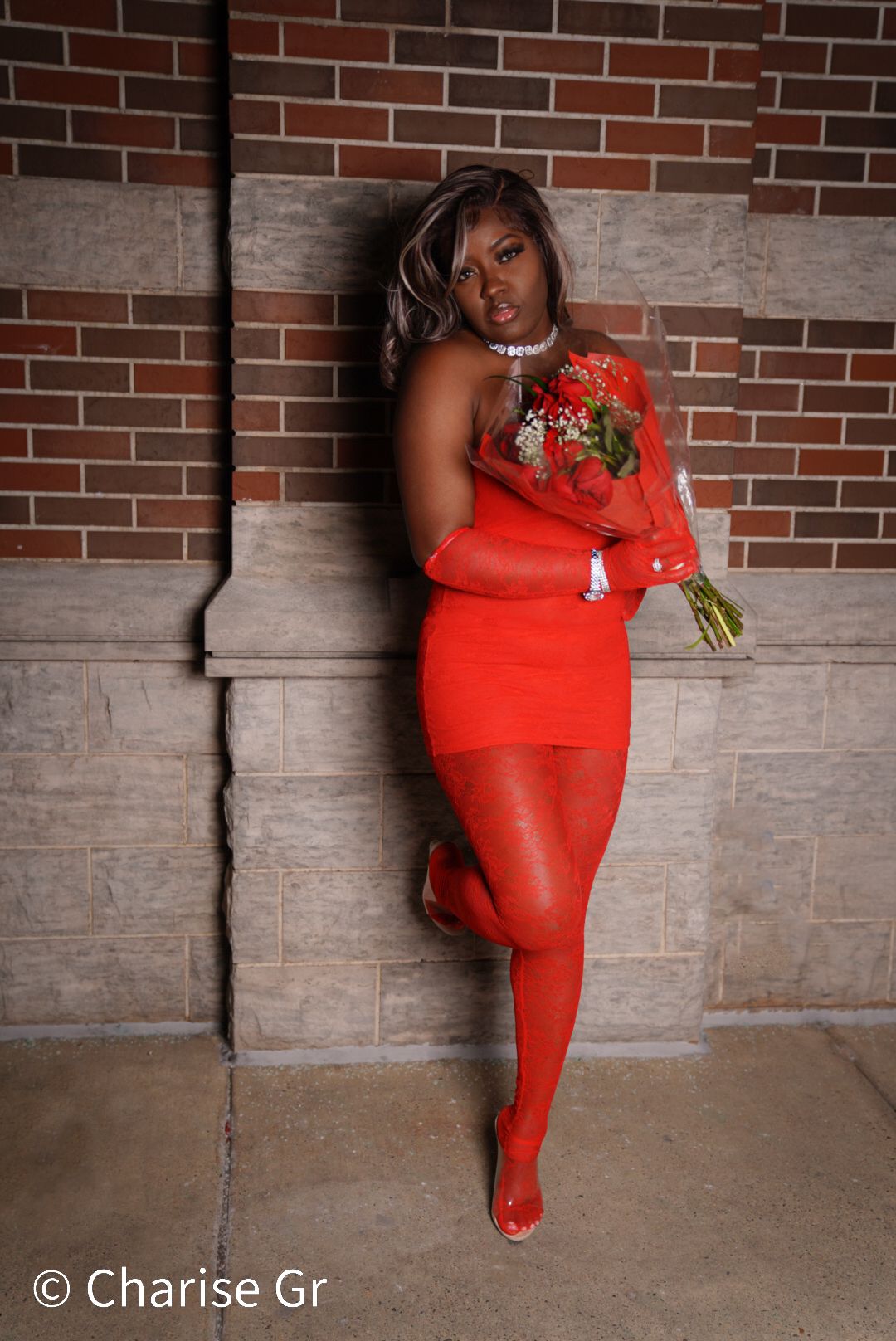 A model in a fitted red dress poses elegantly against a brick wall while holding a bouquet of flowers.