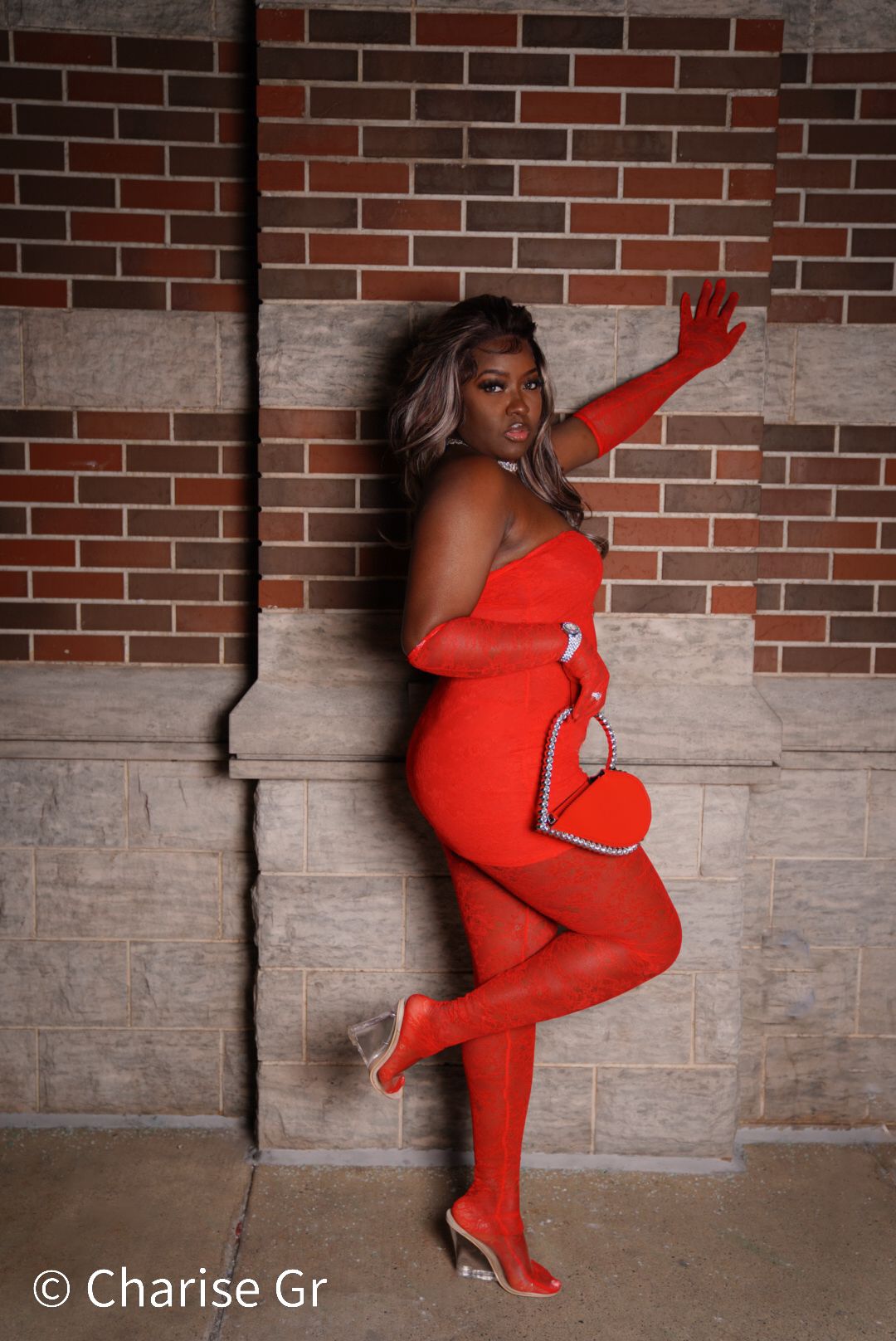 A fashion photoshoot featuring a red bodycon outfit with heart details against a brick and stone wall background.