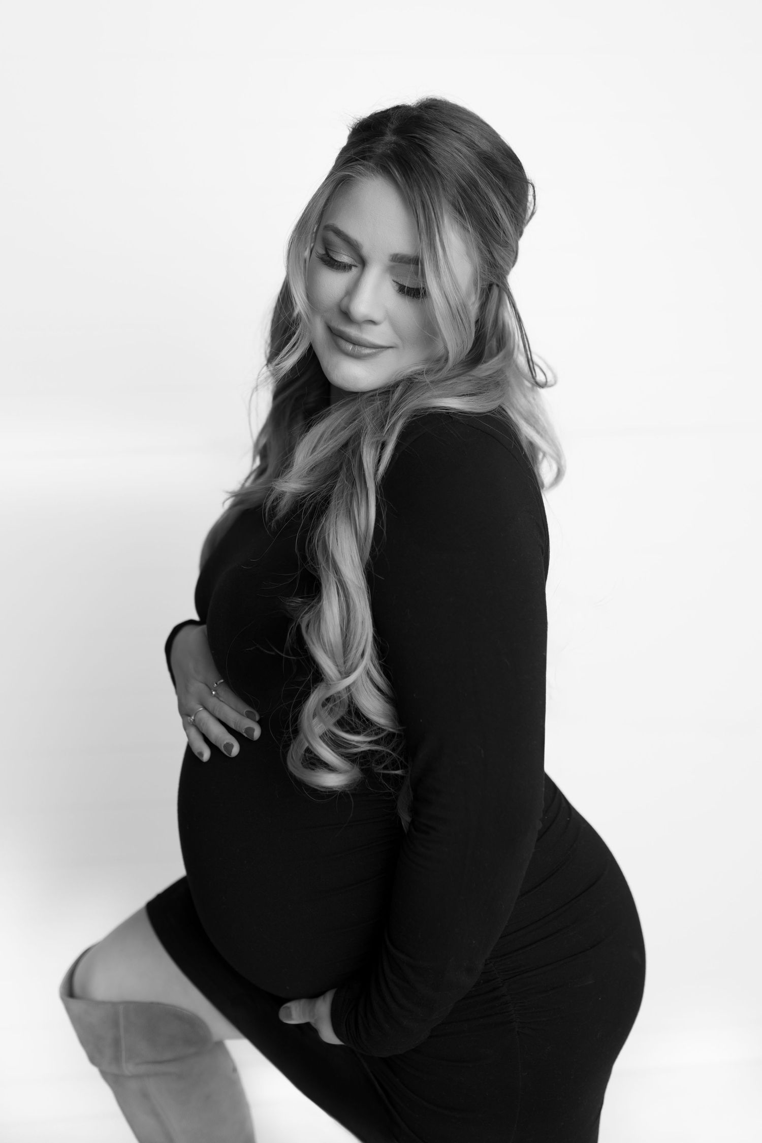 Black and white maternity portrait showing woman in form-fitting long sleeve dress posing elegantly against light background.