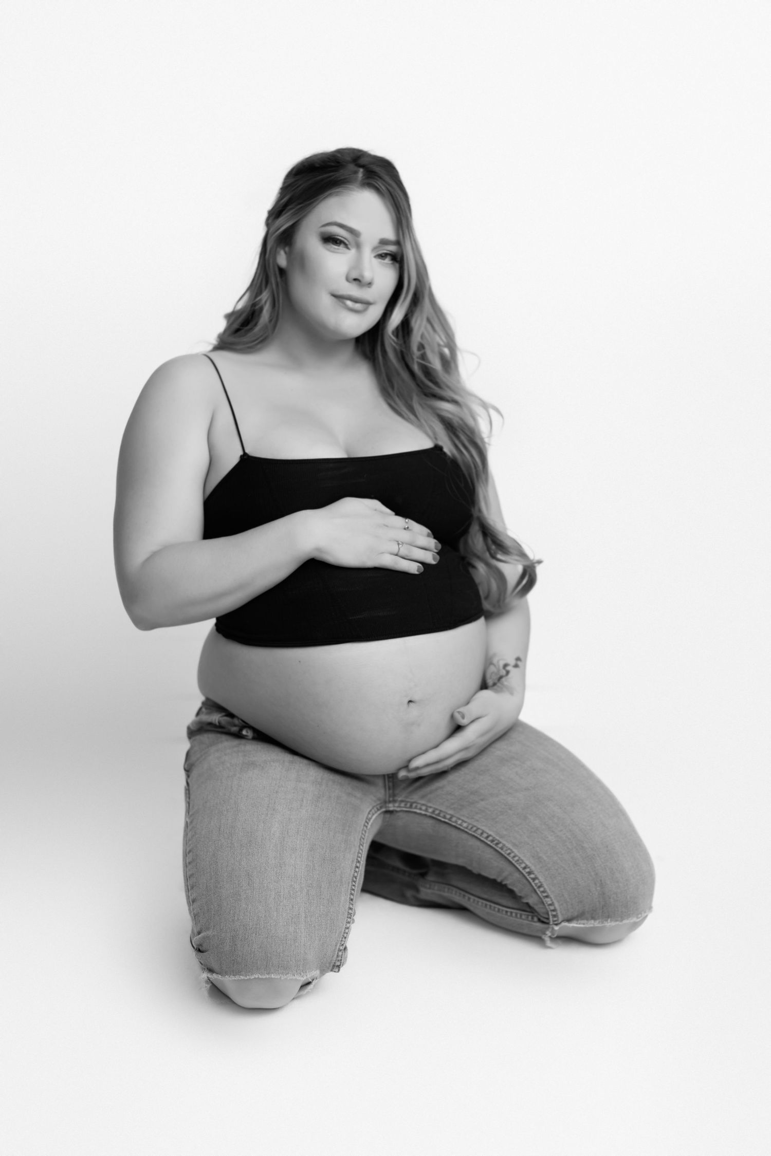 Black and white maternity photo of a pregnant woman in casual attire kneeling against white background.