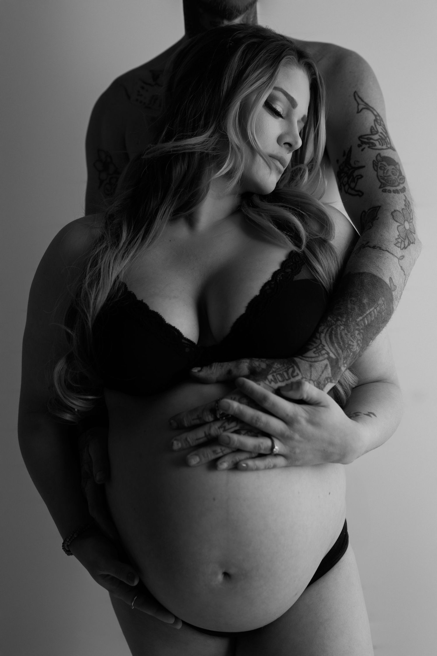 Black and white maternity portrait showing loving embrace with tattooed arms around pregnant belly in intimate pose.