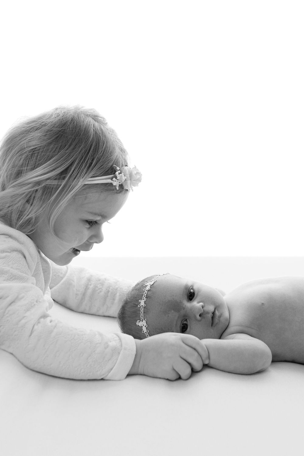 Black and white photo of two young children lying down together, with one wearing a delicate floral headband.