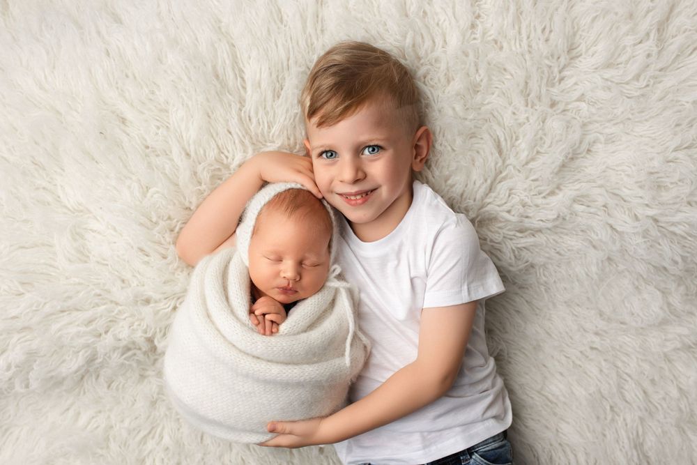 A young child in white shirt poses with a sleeping newborn swaddled in cream-colored wrap on furry white background.