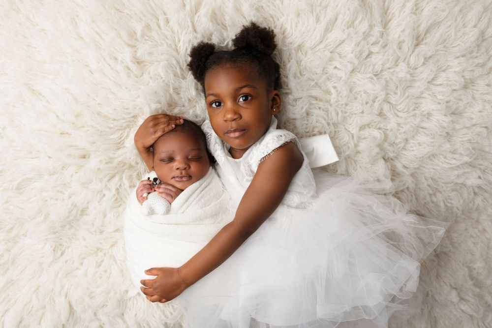 Two children in white dresses lie together on a fluffy white fur background during a portrait session.