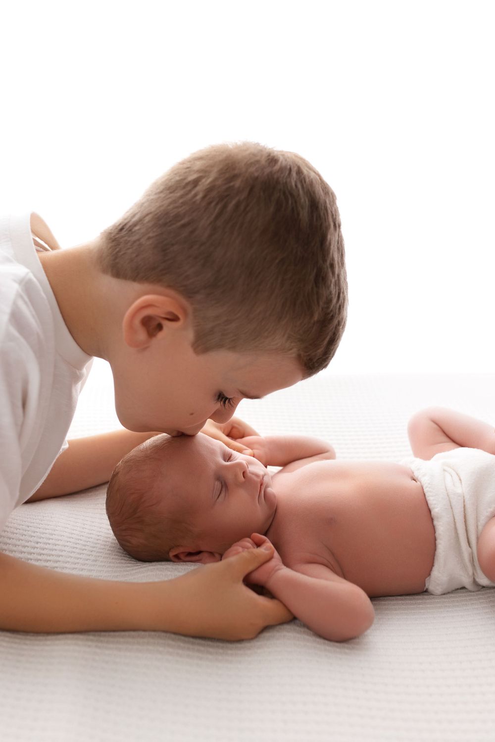 A gentle moment between siblings as an older child leans in close to a baby lying on white bedding.